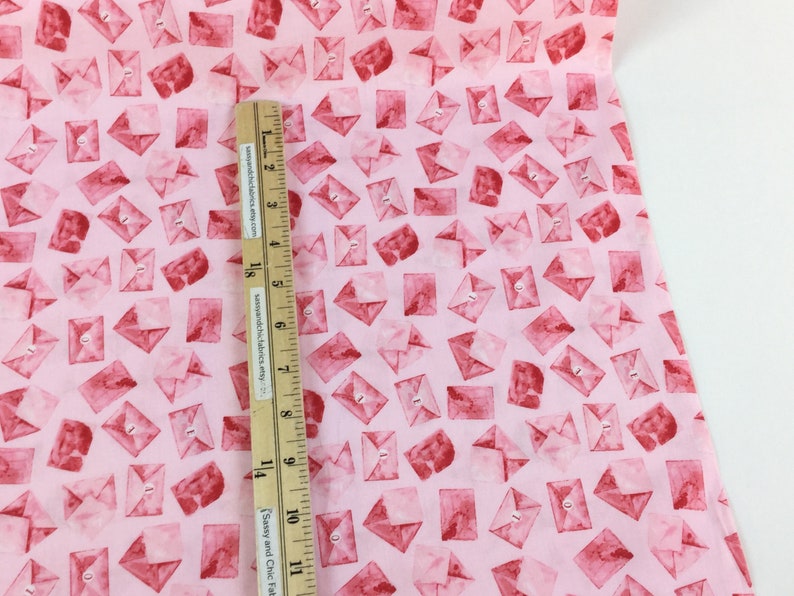 May include: Pink fabric with a repeating pattern of watercolor-style red envelopes. A wooden ruler with inch measurements is placed on the fabric. The fabric is likely for crafting or sewing projects.