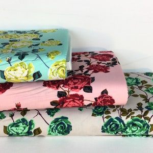 May include: Stacks of floral fabric in various colors. One stack is light blue with yellow roses, another is pink with red roses, and the third is off-white with green roses. The fabric is likely cotton or a similar material, suitable for sewing projects.