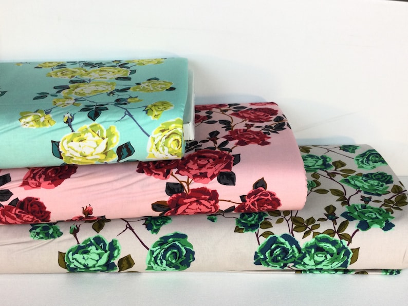 May include: Stacks of floral fabric in various colors. One stack is light blue with yellow roses, another is pink with red roses, and the third is off-white with green roses. The fabric is likely cotton or a similar material, suitable for sewing projects.