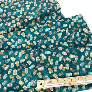 May include: Teal fabric with a repeating pattern of thimbles in various colors. The thimbles have floral and geometric designs. A wooden ruler with inch measurements is in the foreground.