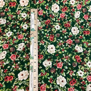 K&ouml;nnte beinhalten: Gr&uuml;ner Stoff mit floralem Muster mit roten und cremefarbenen Rosen, wei&szlig;en Blumen und Tannenzapfen. Ein Lineal ist vertikal auf dem Stoff platziert und zeigt Messungen in Zentimetern. Der Stoff hat ein dichtes, detailliertes Design.