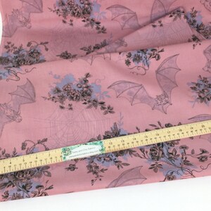 Puede incluir: Tela rosa con un estampado repetido de murci&eacute;lagos, telara&ntilde;as y ramos florales en gris y azul claro. Una regla de madera se coloca en diagonal sobre la tela. La tela es para coser o manualidades.