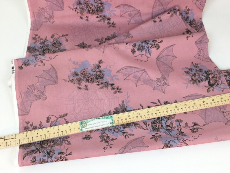 Puede incluir: Tela rosa con un estampado repetido de murci&eacute;lagos, telara&ntilde;as y ramos florales en gris y azul claro. Una regla de madera se coloca en diagonal sobre la tela. La tela es para coser o manualidades.