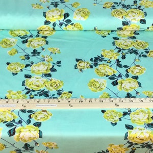 May include: A roll of fabric featuring a repeating floral pattern. The design showcases large yellow roses with green leaves and stems against a light turquoise background. The fabric is printed with the words "Love Always AM".
