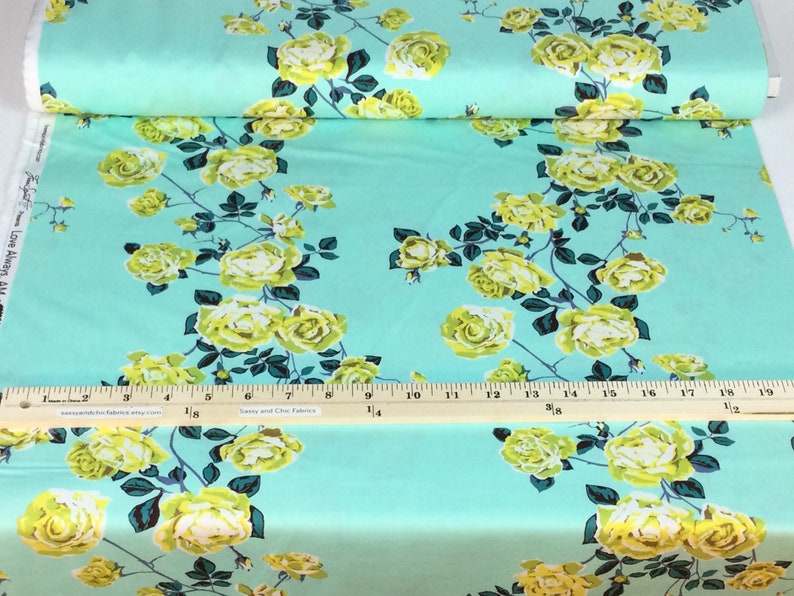 May include: A roll of fabric featuring a repeating floral pattern. The design showcases large yellow roses with green leaves and stems against a light turquoise background. The fabric is printed with the words "Love Always AM".