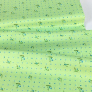 Puede incluir: Una tela verde claro con un patr&oacute;n repetido de bayas azules y hojas, junto con peque&ntilde;os lunares azules. La tela est&aacute; doblada, mostrando su textura y dise&ntilde;o. Adecuada para proyectos de costura y manualidades.