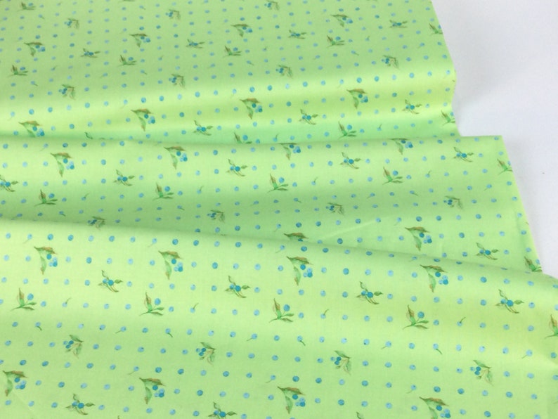 Puede incluir: Una tela verde claro con un patr&oacute;n repetido de bayas azules y hojas, junto con peque&ntilde;os lunares azules. La tela est&aacute; doblada, mostrando su textura y dise&ntilde;o. Adecuada para proyectos de costura y manualidades.