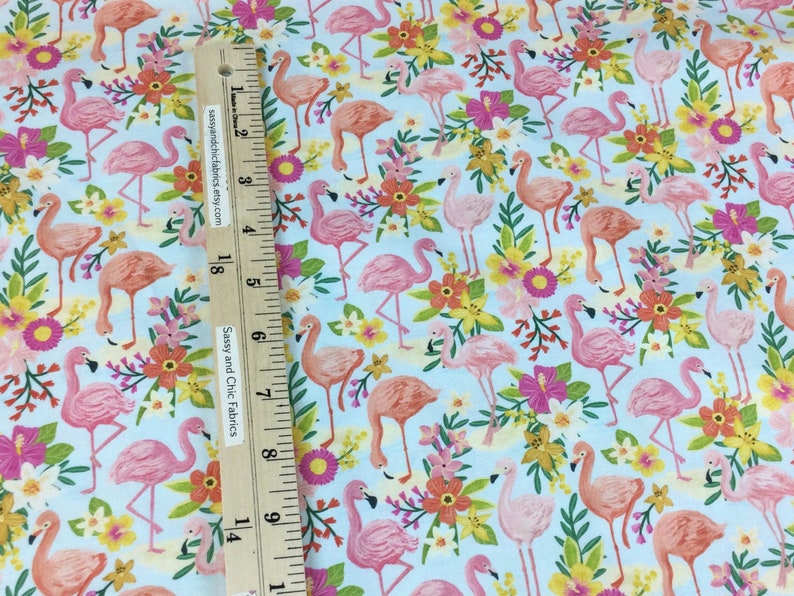 May include: Fabric with a repeating pattern of pink flamingos and colorful flowers on a light blue background. A wooden ruler with the text "Sassy and Chic Fabrics" is in the image.