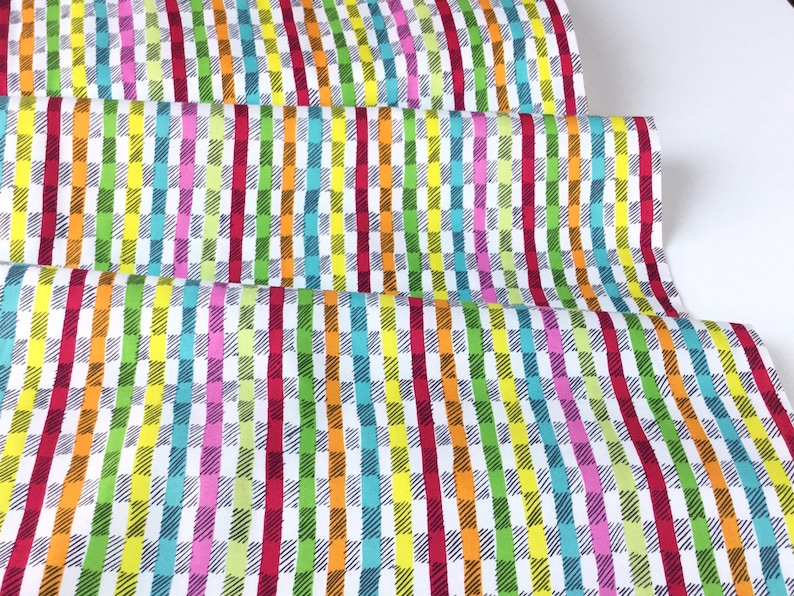 May include: Colorful fabric with a repeating pattern of vertical stripes and checkered blocks. The design features a vibrant mix of colors including red, orange, yellow, green, blue, and pink on a white background. The fabric is folded, showing multiple layers.