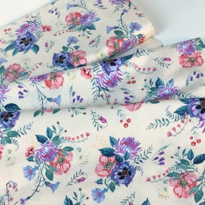 May include: Cream-coloured fabric with a floral design. The pattern includes pink, purple, and blue flowers with green foliage. The fabric is folded, revealing the pattern's repeat across the material.