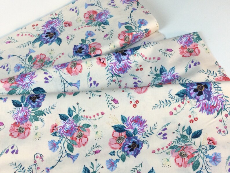 May include: Cream-coloured fabric with a floral design. The pattern includes pink, purple, and blue flowers with green foliage. The fabric is folded, revealing the pattern's repeat across the material.
