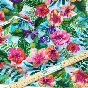 May include: Vibrant fabric featuring a tropical floral pattern. The design showcases large pink, orange, and purple hibiscus flowers and lush green leaves against a light blue background. A wooden ruler is placed on the fabric.