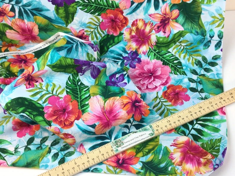 May include: Vibrant fabric featuring a tropical floral pattern. The design showcases large pink, orange, and purple hibiscus flowers and lush green leaves against a light blue background. A wooden ruler is placed on the fabric.