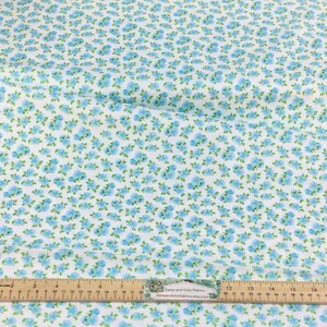 May include: White fabric with a repeating pattern of small blue flowers and green leaves. A wooden ruler with inch markings is placed across the fabric. The fabric is likely used for sewing or crafting projects.