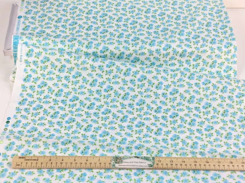 May include: White fabric with a repeating pattern of small blue flowers and green leaves. A wooden ruler with inch markings is placed across the fabric. The fabric is likely used for sewing or crafting projects.