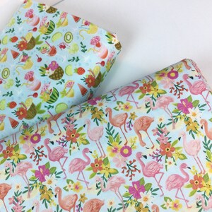 May include: Two rolls of fabric with colorful patterns. One features tropical drinks and fruit on a light blue background. The other has pink flamingos, flowers, and foliage on a light blue background. Both fabrics are rolled up.
