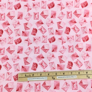 May include: Pink fabric with a repeating pattern of red watercolor envelopes. Each envelope has a small, silver-colored button closure. The fabric is measured with a ruler showing inches.
