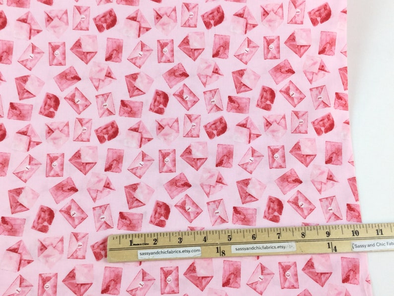 May include: Pink fabric with a repeating pattern of red watercolor envelopes. Each envelope has a small, silver-colored button closure. The fabric is measured with a ruler showing inches.