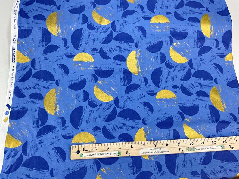 May include: Blue fabric with a pattern of gold and navy blue half-circles and brushstrokes. A wooden ruler with inch markings is placed on the fabric. The fabric has the text "Robert Kaufman" and "SassyandChicFabrics.etsy.com".