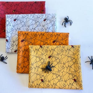 May include: Four folded fabric squares in red, white, orange, and yellow, each with a black spiderweb pattern and small red spiders. Black plastic spiders are scattered around the squares, creating a Halloween theme.