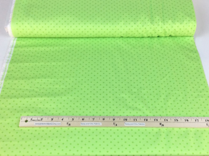 May include: Bright green fabric with small, evenly spaced, darker green polka dots. A wooden ruler with inch measurements is placed on the fabric. The fabric is rolled up in the background. The website address "sassyandchicfabrics.etsy.com" is printed on the ruler.
