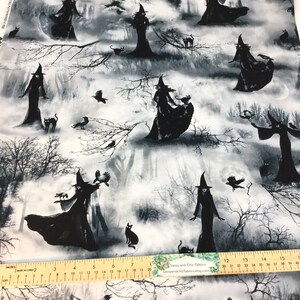 May include: Fabric with a black and white Halloween design. Silhouetted witches in tall hats and flowing gowns are scattered across a misty forest scene. Black cats and crows are also featured. The fabric is printed on a white background.