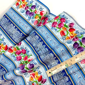 May include: Fabric with a repeating pattern of colorful tulips and Dutch houses on a light blue background. The design includes vertical stripes and floral accents, creating a vibrant and decorative textile.
