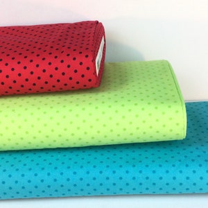 May include: Three rolls of fabric in red, green, and blue, each with a polka dot pattern. The red fabric has black dots, the green has lighter green dots, and the blue has darker blue dots. The fabrics are stacked on top of each other.