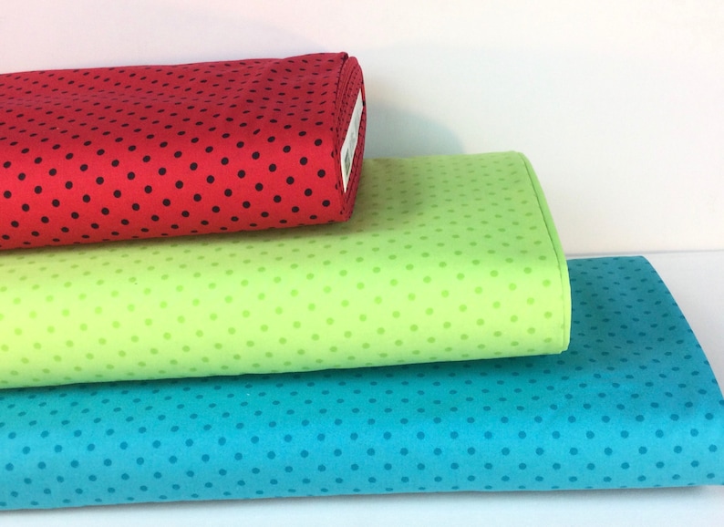 May include: Three rolls of fabric in red, green, and blue, each with a polka dot pattern. The red fabric has black dots, the green has lighter green dots, and the blue has darker blue dots. The fabrics are stacked on top of each other.