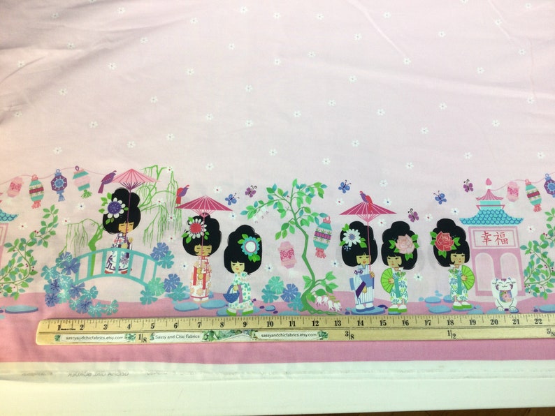 May include: Pink fabric with a repeating border design featuring stylized Japanese dolls, pagodas, lanterns, and butterflies. The dolls wear floral hairpieces and hold parasols. The fabric has a light pink background with small white floral accents.