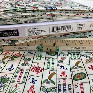 May include: A roll of cotton fabric with a Mahjong tile pattern in red, green, and blue on an off-white background. The fabric is labeled "Timeless Treasures" and "100% Cotton". A wooden ruler is placed on top of the fabric.