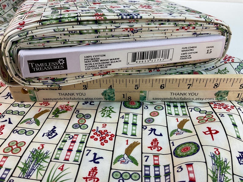 May include: A roll of cotton fabric with a Mahjong tile pattern in red, green, and blue on an off-white background. The fabric is labeled "Timeless Treasures" and "100% Cotton". A wooden ruler is placed on top of the fabric.