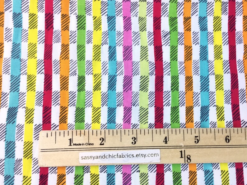 May include: Colorful fabric with a repeating pattern of vertical stripes in red, orange, yellow, green, blue, and pink. Each stripe is overlaid with a black and white checkered design. A wooden ruler shows measurements in inches.
