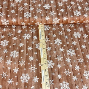 Pu&ograve; includere: Tessuto marrone con motivo a venature del legno e fiocchi di neve bianchi. Il tessuto &egrave; piegato, mostrando il motivo sul davanti e il bordo bianco. Un righello &egrave; posto sul tessuto.