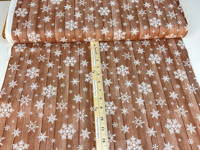 Pu&ograve; includere: Tessuto marrone con motivo a venature del legno e fiocchi di neve bianchi. Il tessuto &egrave; piegato, mostrando il motivo sul davanti e il bordo bianco. Un righello &egrave; posto sul tessuto.