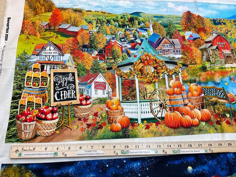 Op de afbeelding: Stoffpaneel met een herfsttafereel met een prieel, pompoenen en een bord voor "Apple Cider". Het kunstwerk toont een schilderachtig dorp met kleurrijk gebladerte, vaten met appels en een watermolen. Een liniaal toont inches.