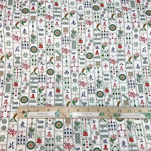 May include: Mahjong tile fabric with a repeating pattern of tiles, flowers, and Chinese characters in red, green, and black on a cream background. A wooden ruler with inches is placed on top of the fabric.