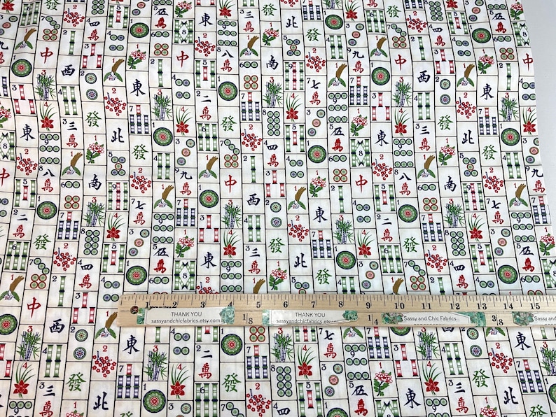 May include: Mahjong tile fabric with a repeating pattern of tiles, flowers, and Chinese characters in red, green, and black on a cream background. A wooden ruler with inches is placed on top of the fabric.