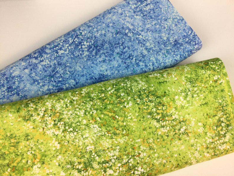 May include: Two rolls of fabric, one in a light blue with white speckles, the other in a vibrant green with white and yellow floral patterns. The fabrics appear to be cotton or a similar material, suitable for quilting or crafting projects.