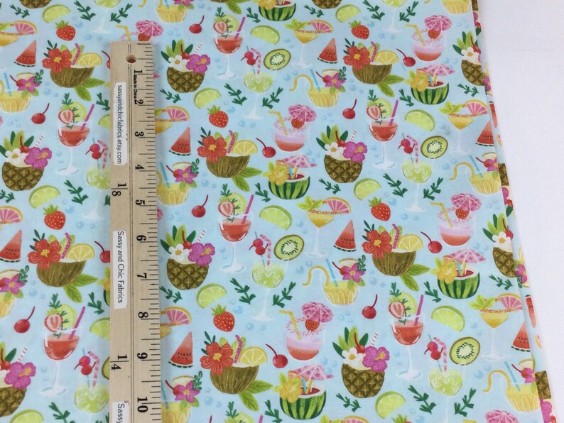 May include: Light blue fabric with a tropical cocktail pattern. The design includes cocktails, pineapples, watermelon, and flowers. A wooden ruler with text is placed on the fabric. The text on the ruler says "Sassy and Chic Fabrics etsy.com".