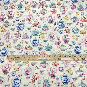 May include: Cream-colored fabric with a repeating pattern of teapots, teacups, and floral arrangements in shades of blue, pink, and green. A wooden ruler with inch markings is placed across the fabric.