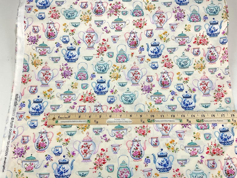 May include: Cream-colored fabric with a repeating pattern of teapots, teacups, and floral arrangements in shades of blue, pink, and green. A wooden ruler with inch markings is placed across the fabric.