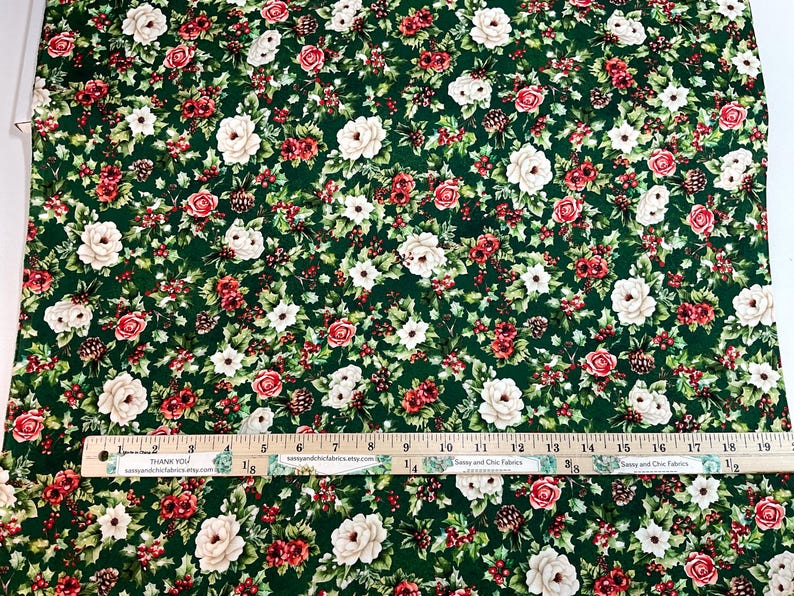 K&ouml;nnte beinhalten: Gr&uuml;ner Stoff mit einem Blumenmuster mit roten und wei&szlig;en Rosen, kleinen wei&szlig;en Blumen und roten Beeren. Ein Holzlineal mit Zentimetermessungen liegt auf dem Stoff. Der Stoff hat einen dunkelgr&uuml;nen Hintergrund.