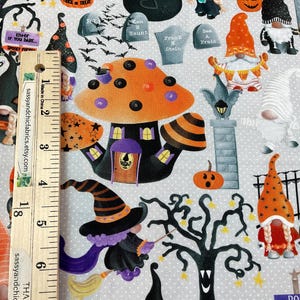 May include: Halloween-themed fabric featuring a whimsical design. The pattern includes orange pumpkins, witch hats, gnomes, a haunted mushroom house, tombstones, and bats. The fabric has a light gray background with white polka dots. A wooden ruler is placed on the fabric.