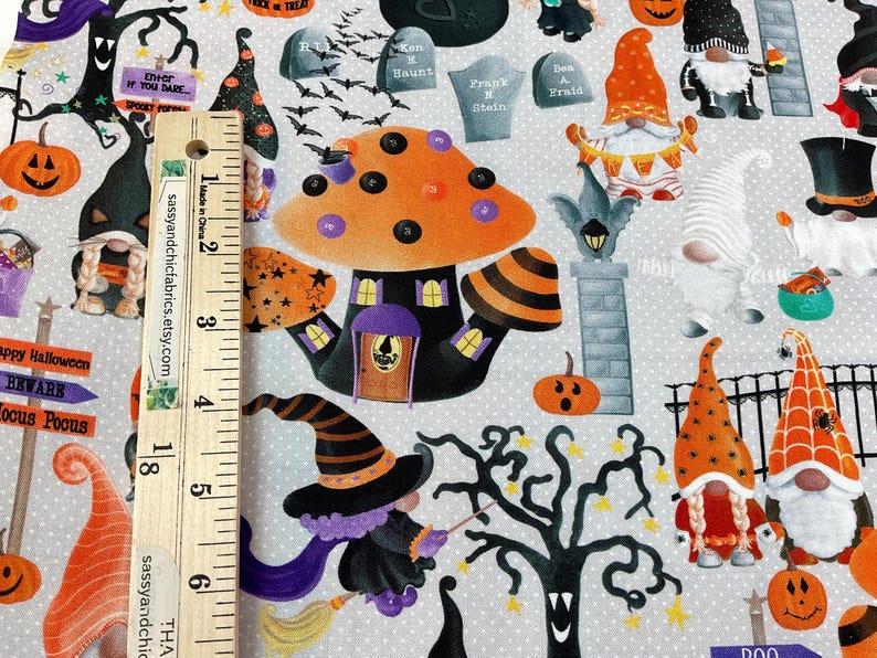 May include: Halloween-themed fabric featuring a whimsical design. The pattern includes orange pumpkins, witch hats, gnomes, a haunted mushroom house, tombstones, and bats. The fabric has a light gray background with white polka dots. A wooden ruler is placed on the fabric.