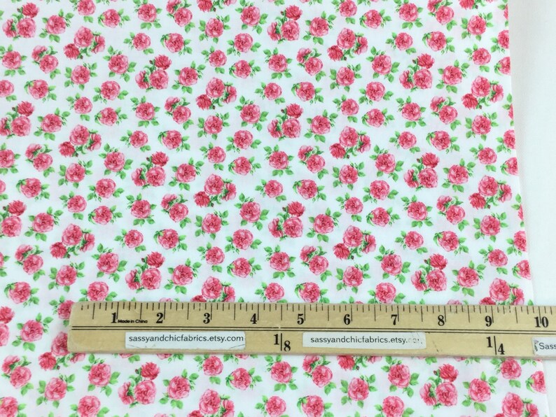 May include: White fabric covered in a repeating pattern of pink roses with green leaves. A ruler with inch markings is along the bottom. The text "sassyandchicfabrics.etsy.com" is printed on the ruler.