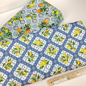 May include: Two rolls of fabric with fruit patterns. One roll features lemons and oranges on a blue and white tile design. The other roll has a pattern of lemons on a white and blue diamond design. A wooden ruler is in the foreground.