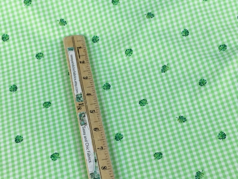 K&ouml;nnte beinhalten: Gr&uuml;ner und wei&szlig;er Vichystoff mit kleinen gr&uuml;nen Blattmustern. Ein Holzlineal mit dem Text "Sassy and Chic Fabrics" liegt diagonal &uuml;ber dem Stoff und misst in Zentimetern.