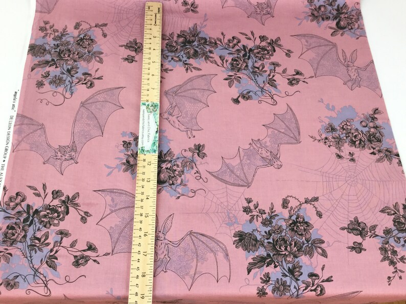 Puede incluir: Tela rosa con un patr&oacute;n repetido de murci&eacute;lagos, flores y telara&ntilde;as. Los murci&eacute;lagos est&aacute;n en vuelo y las flores est&aacute;n agrupadas. Una regla de madera se coloca verticalmente sobre la tela.