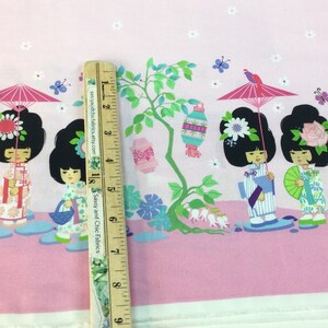 May include: Pink fabric with a repeating pattern of cartoon geishas, parasols, and lanterns. The geishas are in traditional attire, with black hair and floral accents. The fabric is measured with a wooden ruler.